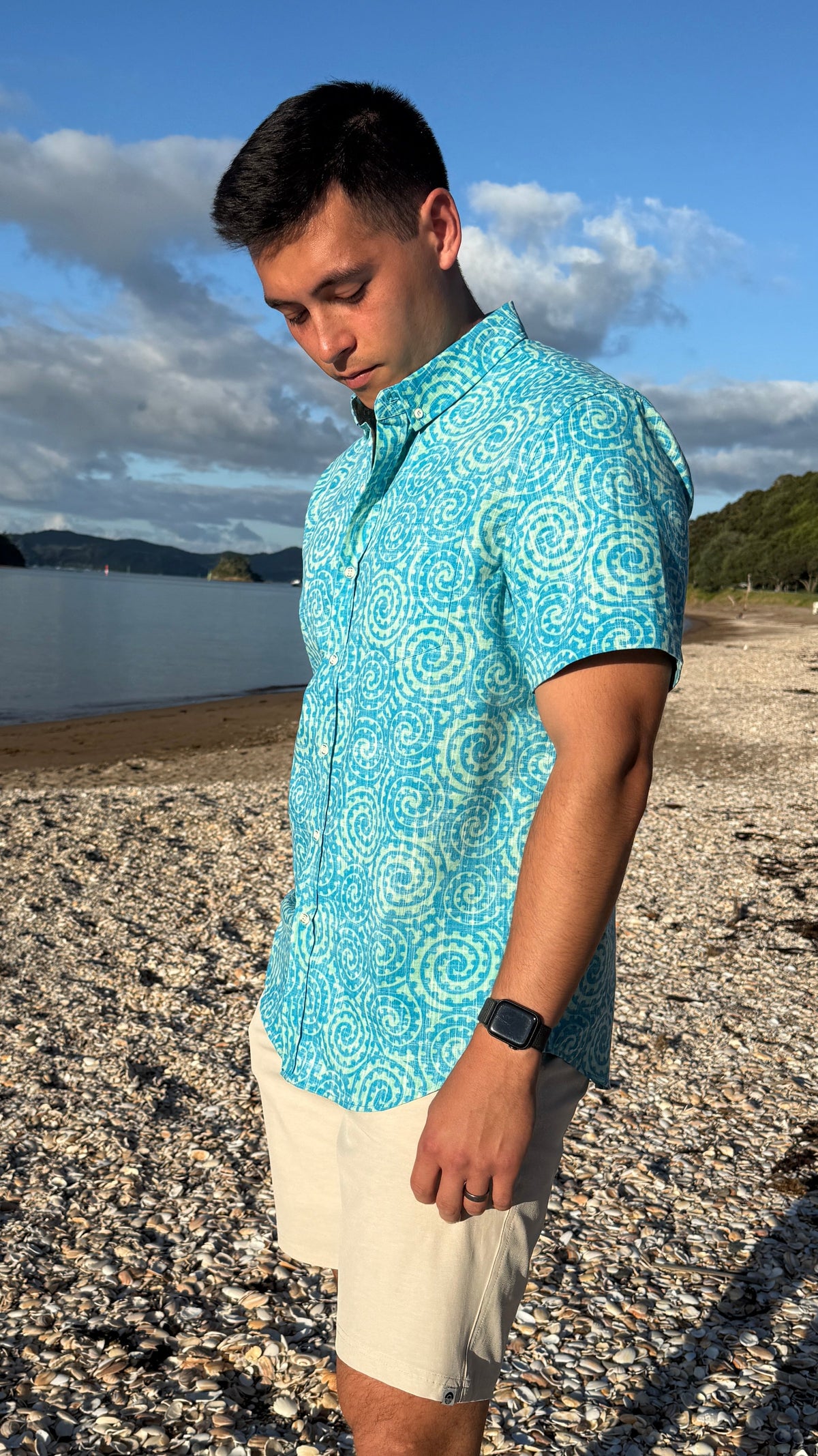 A man wearing a turquoise and white patterned short-sleeve button-up shirt and beige shorts, standing on a pebble beach with water and hills in the background.