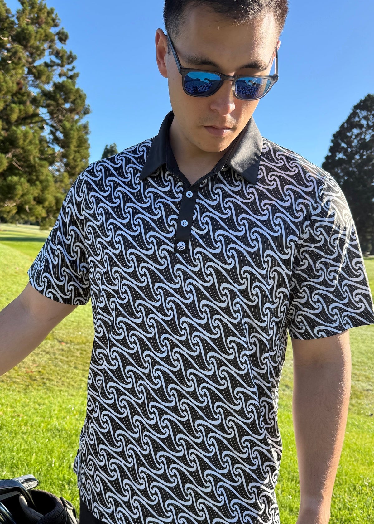 Kaleo Pūhoro men’s golf polo with a flowing white print on black, short sleeves, a black collar and three-button placket, worn by a model on a golf course.