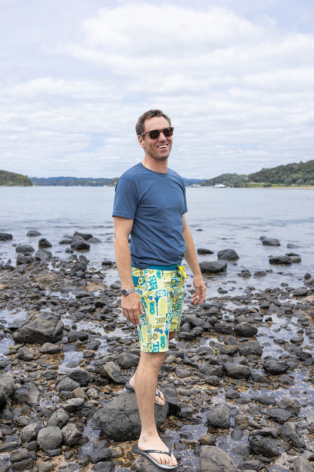 Men’s Tiki boardshorts in bold green, aqua, and yellow Polynesian-inspired design with manaia and tiki motifs. Lightweight quick-dry swim shorts, perfect for summer beachwear, surfing, and water sports in New Zealand. Features back pocket with secure closure, drawstring waist, and durable fabric for comfort and performance.