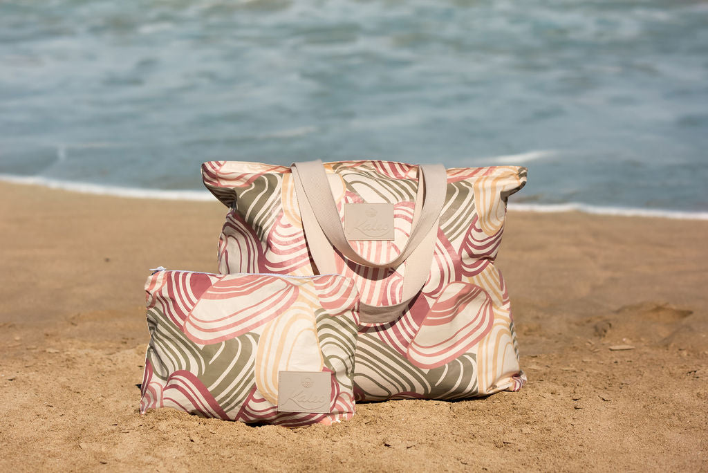 Woman holding a Pipi Shell splashproof tote bag on a New Zealand beach. Large, lightweight, and water-resistant tote with coastal shell print design. Perfect for carrying towels, swimwear, or beach essentials while keeping belongings dry. Stylish Kiwi summer beach bag for holidays, poolside, and travel.