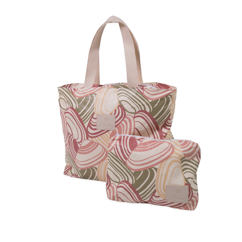 Woman holding a Pipi Shell splashproof tote bag on a New Zealand beach. Large, lightweight, and water-resistant tote with coastal shell print design. Perfect for carrying towels, swimwear, or beach essentials while keeping belongings dry. Stylish Kiwi summer beach bag for holidays, poolside, and travel.