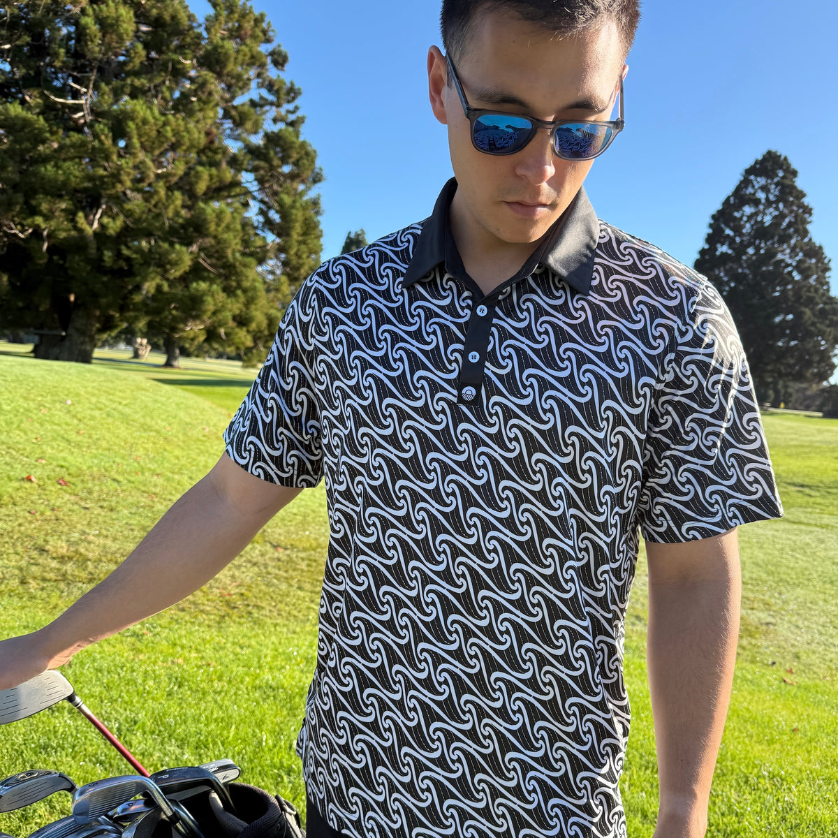 Kaleo Pūhoro men’s golf polo with a flowing white print on black, short sleeves, a black collar and three-button placket, worn by a model on a golf course.