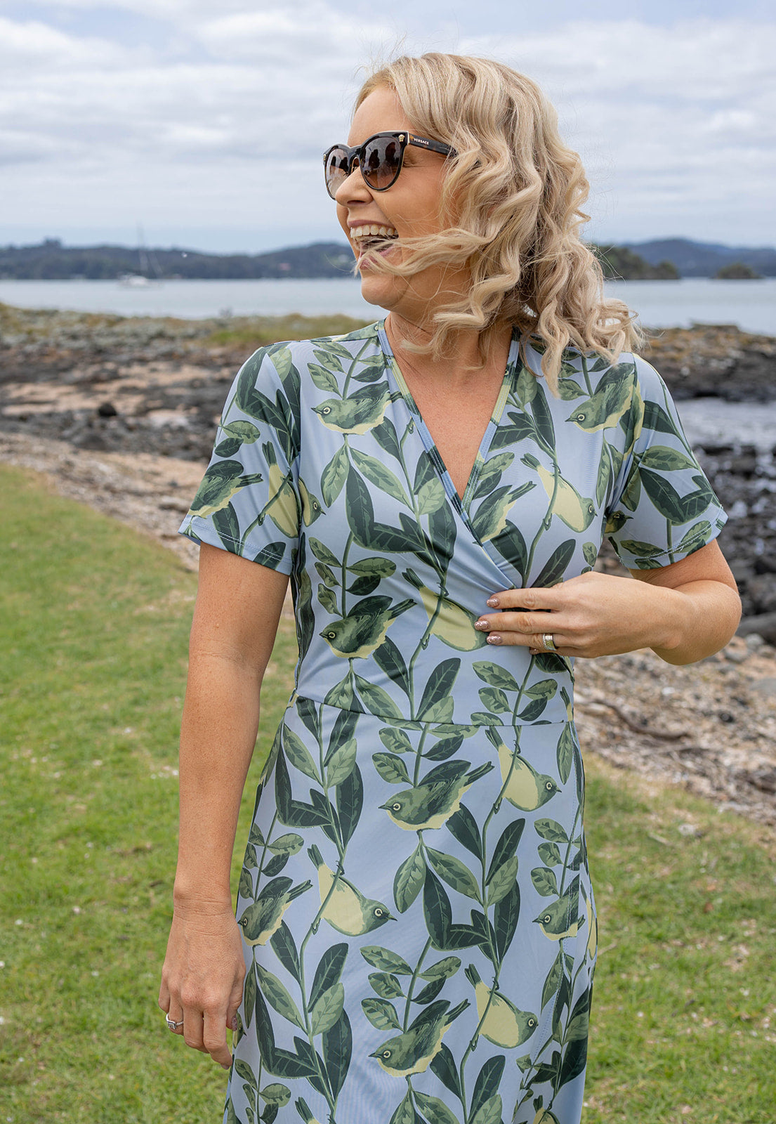 Elegant Tauhou / Wax-Eye wrap dress featuring a soft blue base with green wax-eye bird and foliage print. New Zealand-inspired women’s dress with flattering wrap design, short sleeves, and knee-length cut. Perfect for summer outings, coastal holidays, and everyday Kiwi style.