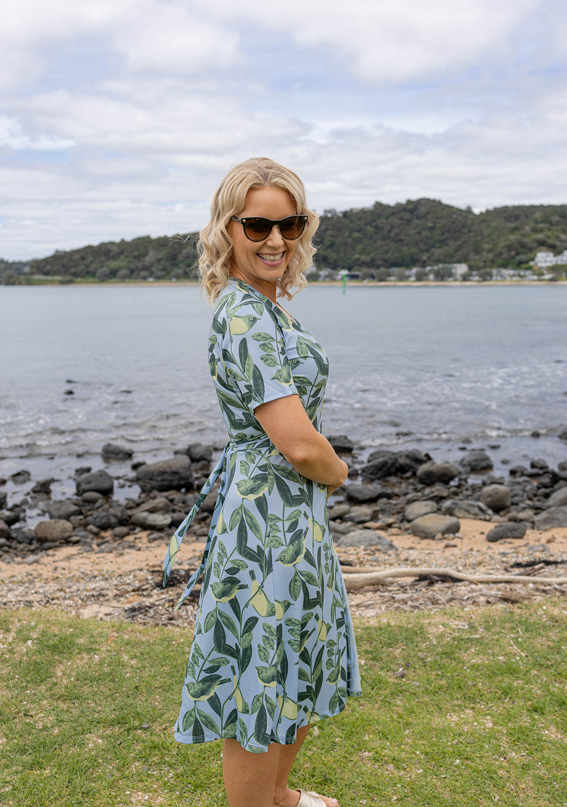 Elegant Tauhou / Wax-Eye wrap dress featuring a soft blue base with green wax-eye bird and foliage print. New Zealand-inspired women’s dress with flattering wrap design, short sleeves, and knee-length cut. Perfect for summer outings, coastal holidays, and everyday Kiwi style.