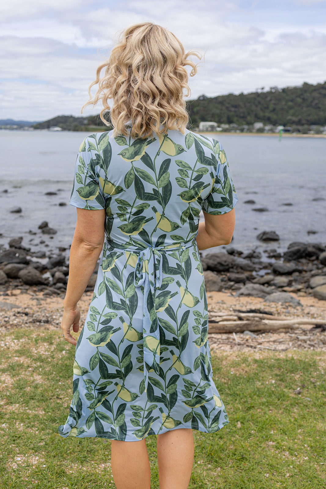 Elegant Tauhou / Wax-Eye wrap dress featuring a soft blue base with green wax-eye bird and foliage print. New Zealand-inspired women’s dress with flattering wrap design, short sleeves, and knee-length cut. Perfect for summer outings, coastal holidays, and everyday Kiwi style.