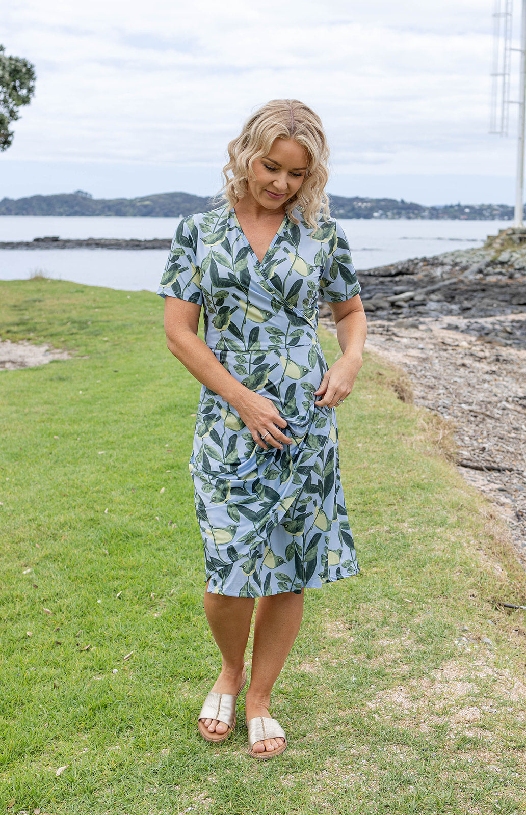 Elegant Tauhou / Wax-Eye wrap dress featuring a soft blue base with green wax-eye bird and foliage print. New Zealand-inspired women’s dress with flattering wrap design, short sleeves, and knee-length cut. Perfect for summer outings, coastal holidays, and everyday Kiwi style.