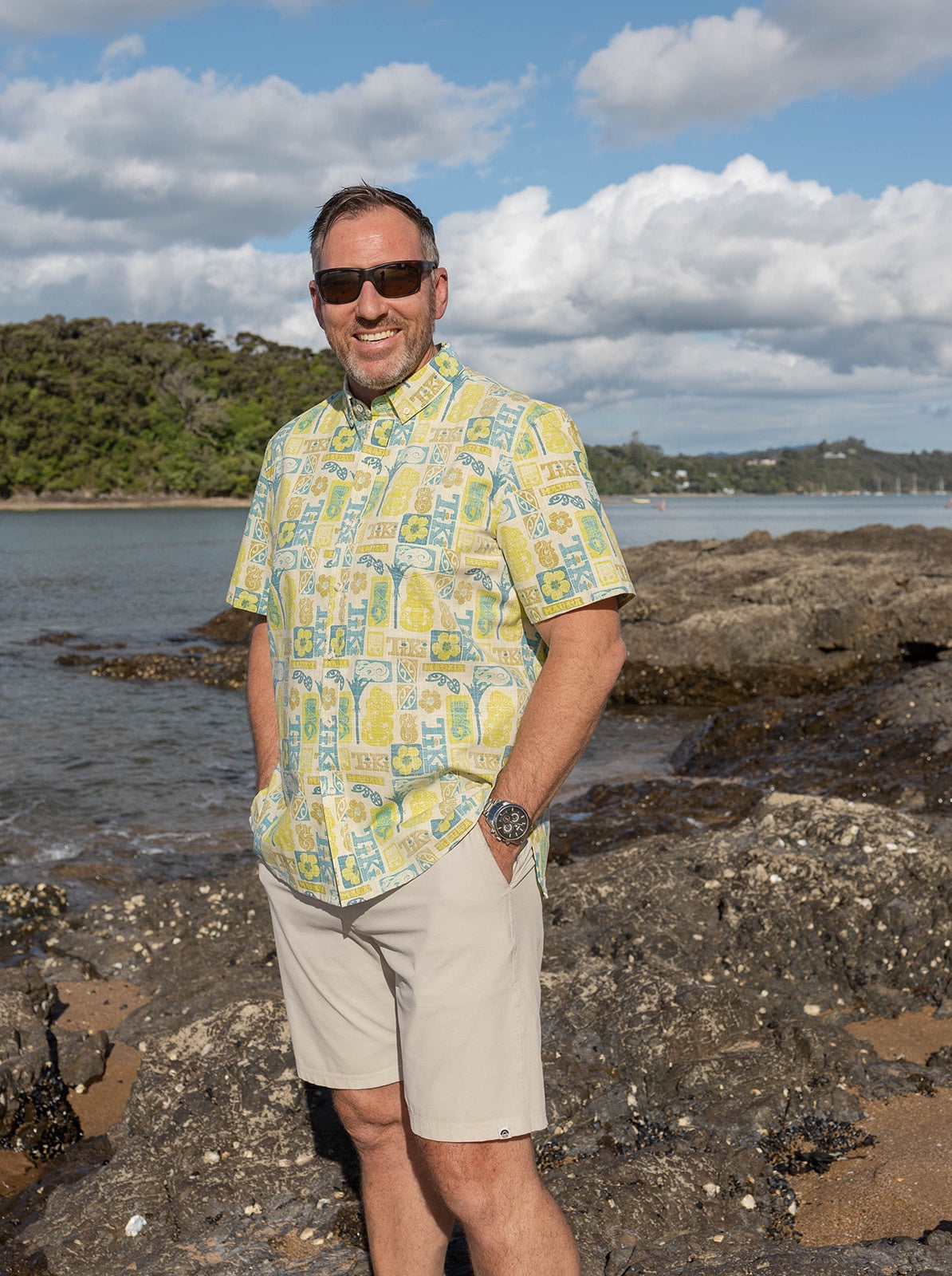 Men’s Tiki Manaia Kiwi Classic short sleeve shirt in vibrant green, blue, and yellow Polynesian-inspired print. Lightweight cotton blend perfect for summer, beach holidays, and New Zealand resort wear. Features bold tiki, manaia, and floral motifs for a unique island style.