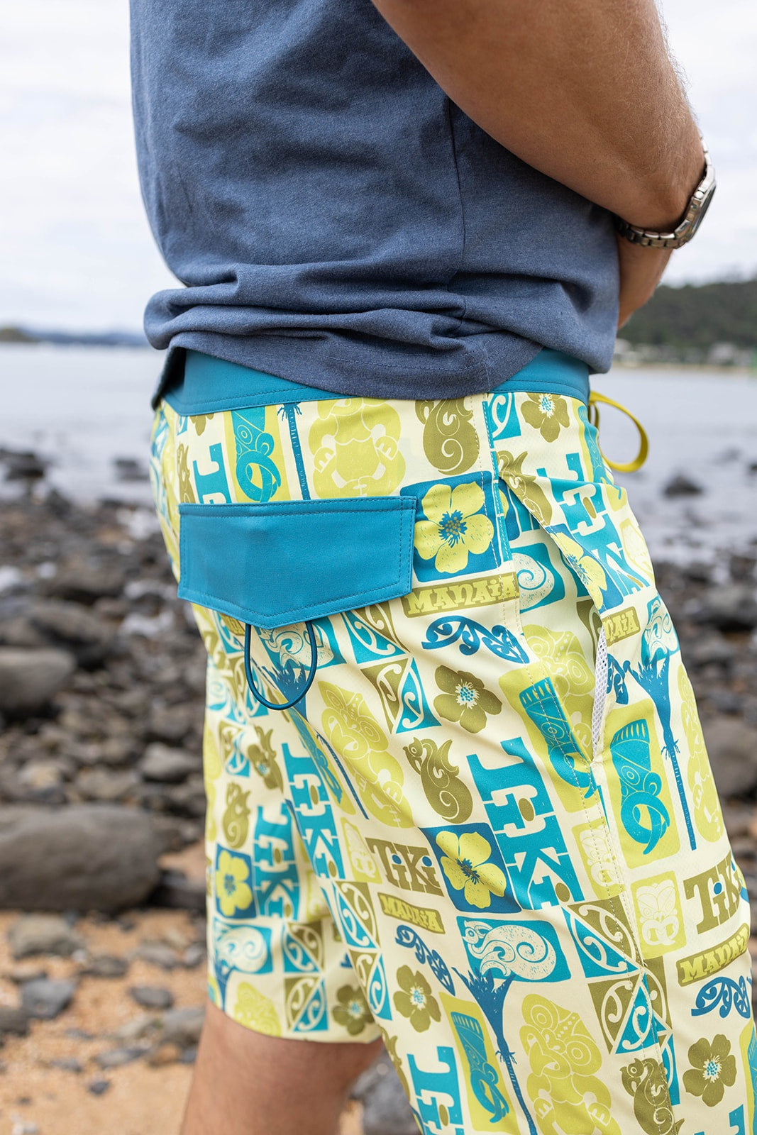 Men’s Tiki boardshorts in bold green, aqua, and yellow Polynesian-inspired design with manaia and tiki motifs. Lightweight quick-dry swim shorts, perfect for summer beachwear, surfing, and water sports in New Zealand. Features back pocket with secure closure, drawstring waist, and durable fabric for comfort and performance.